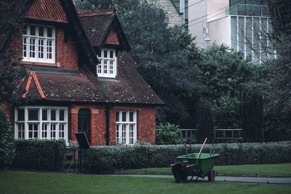 A well-maintained garden in front of a red brick house with white-framed windows and a brown tiled roof. The garden features trimmed hedges and a lush green lawn, with a hedge separating the lawn from